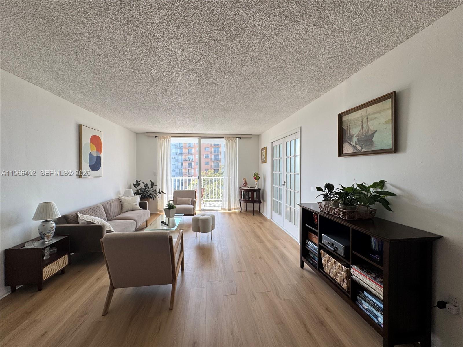 A living room features light wood flooring, a textured ceiling, and a balcony with city views — 1470 NE 125th Ter # 407, North Miami FL 33161, NORTH MIAMI, FL