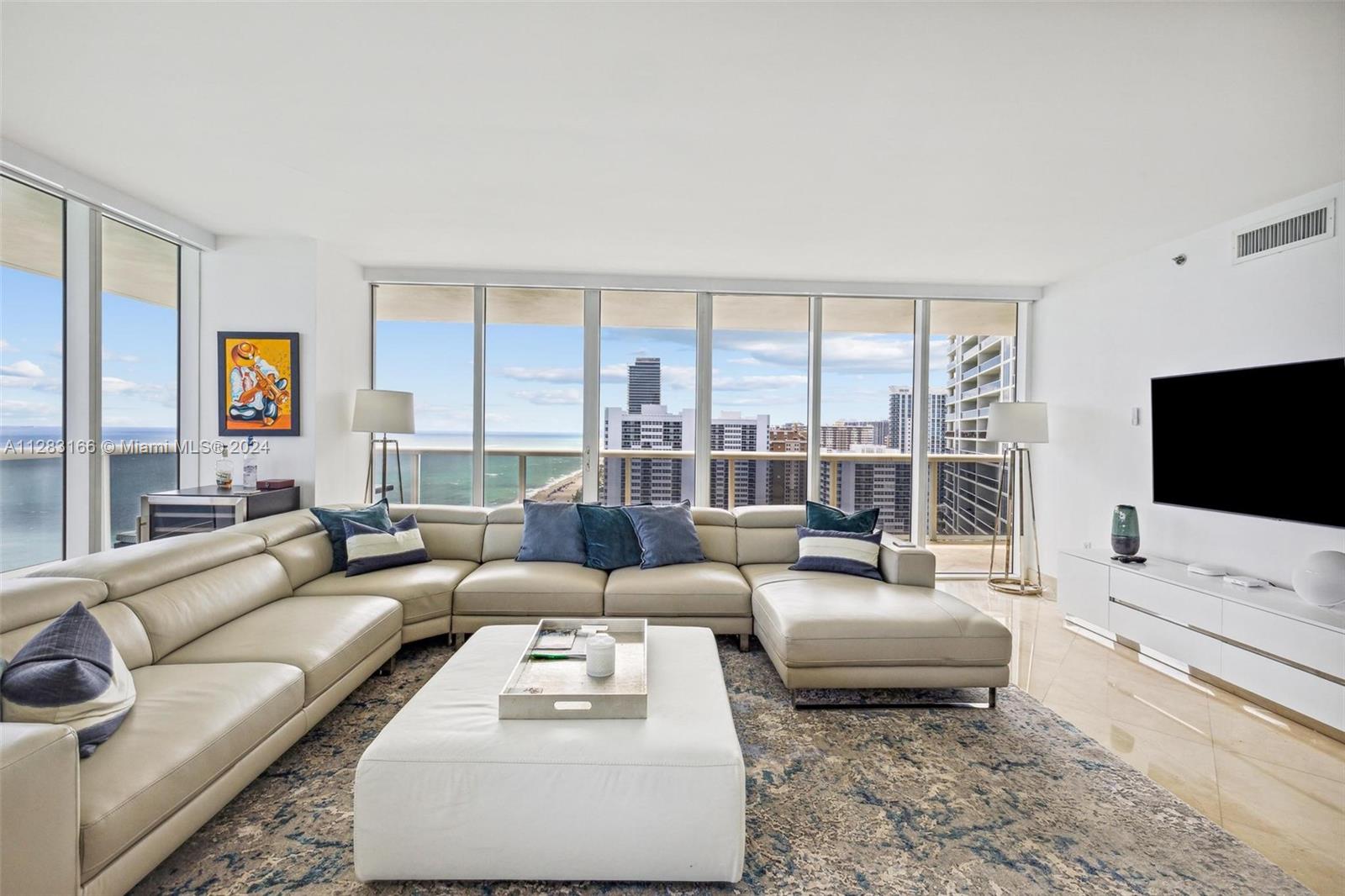 A modern living room features a large beige sectional sofa, white ottoman, and floor-to-ceiling windows — 1830 S OCEAN DR # 2102, Hallandale Beach FL 33009, HALLANDALE BEACH, FL