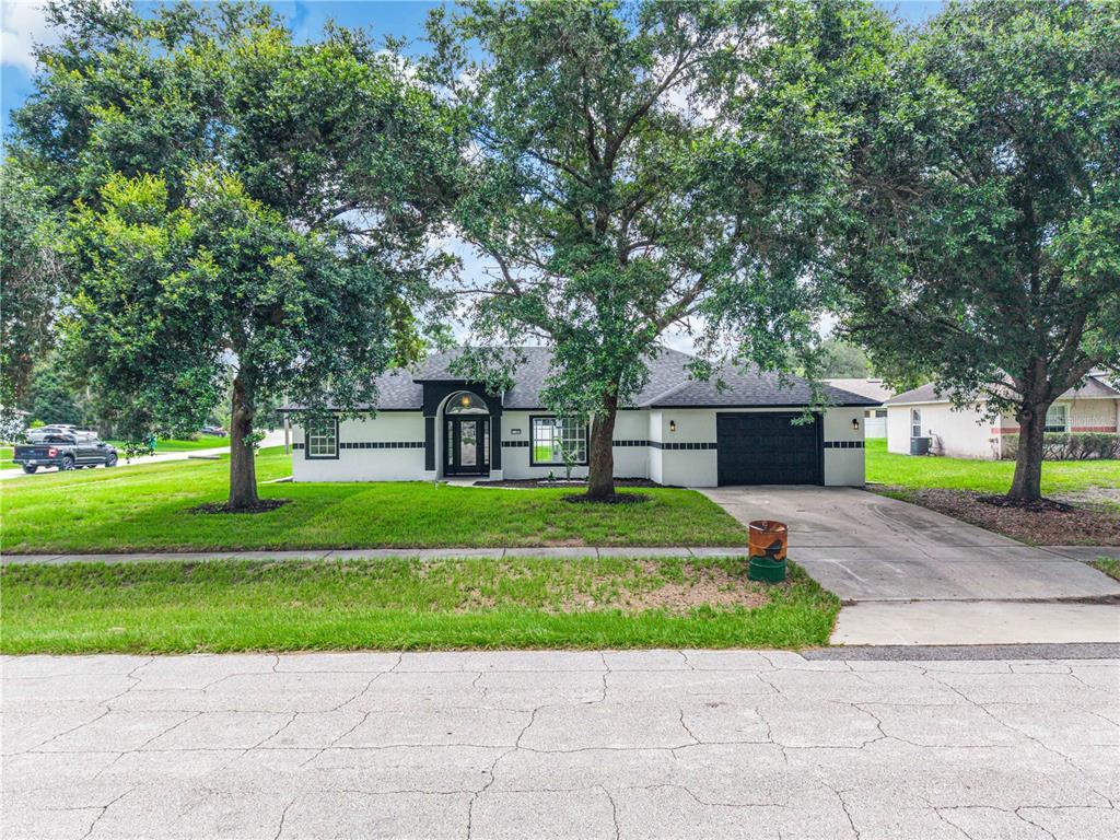 A single-story white house with black trim and a two-car garage sits behind large trees — 2570 COACHMAN DRIVE, DELTONA, FL