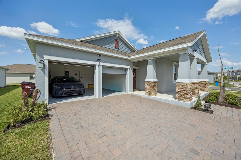 A gray house with a two-car garage, stone-accented porch columns, and a paver driveway — 2466 RELAXING LANE, KISSIMMEE, FL