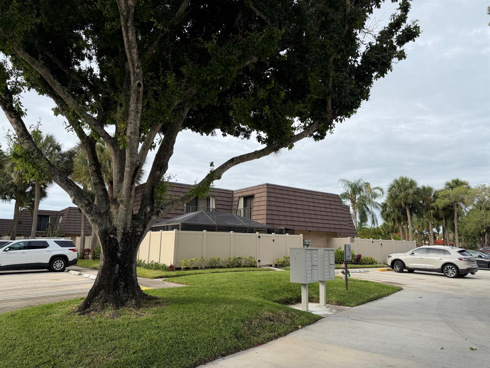 A large tree stands in front of a multi-story apartment building with a fenced patio — 7622 N 76th Way # 200, West Palm Beach FL 33407, WEST PALM BEACH, FL