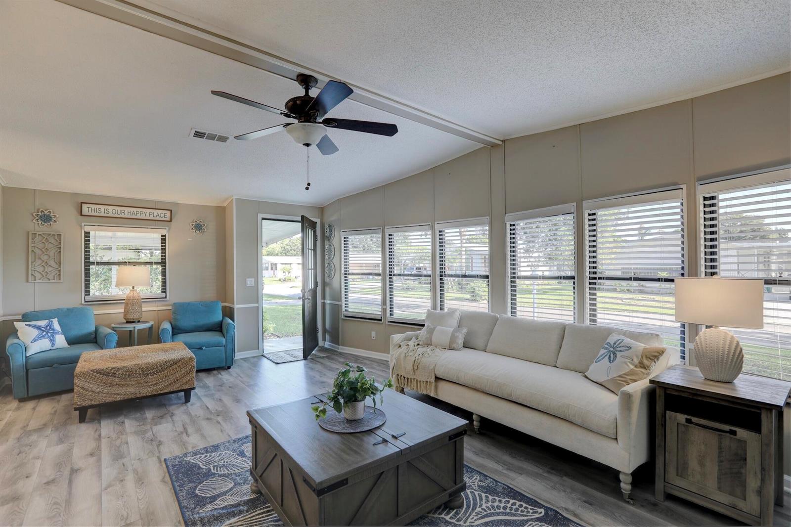 A living room features light wood flooring, a white sofa, blue armchairs, and large windows — BAREFOOT BAY, FL