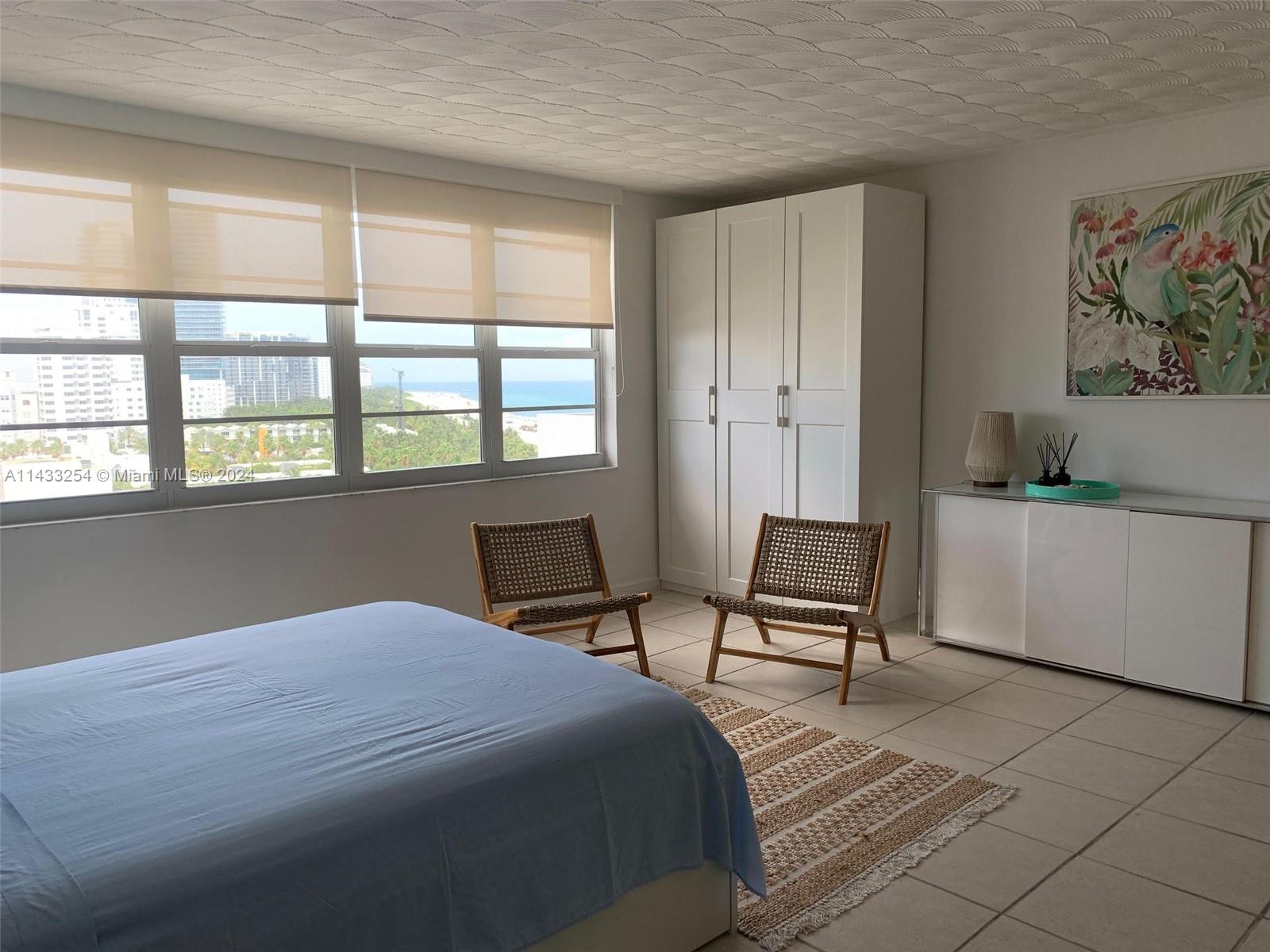 A bedroom features a blue bed, two woven chairs, a white wardrobe, and ocean views — 100 Lincoln Rd # 1039, Miami Beach FL 33139, MIAMI BEACH, FL
