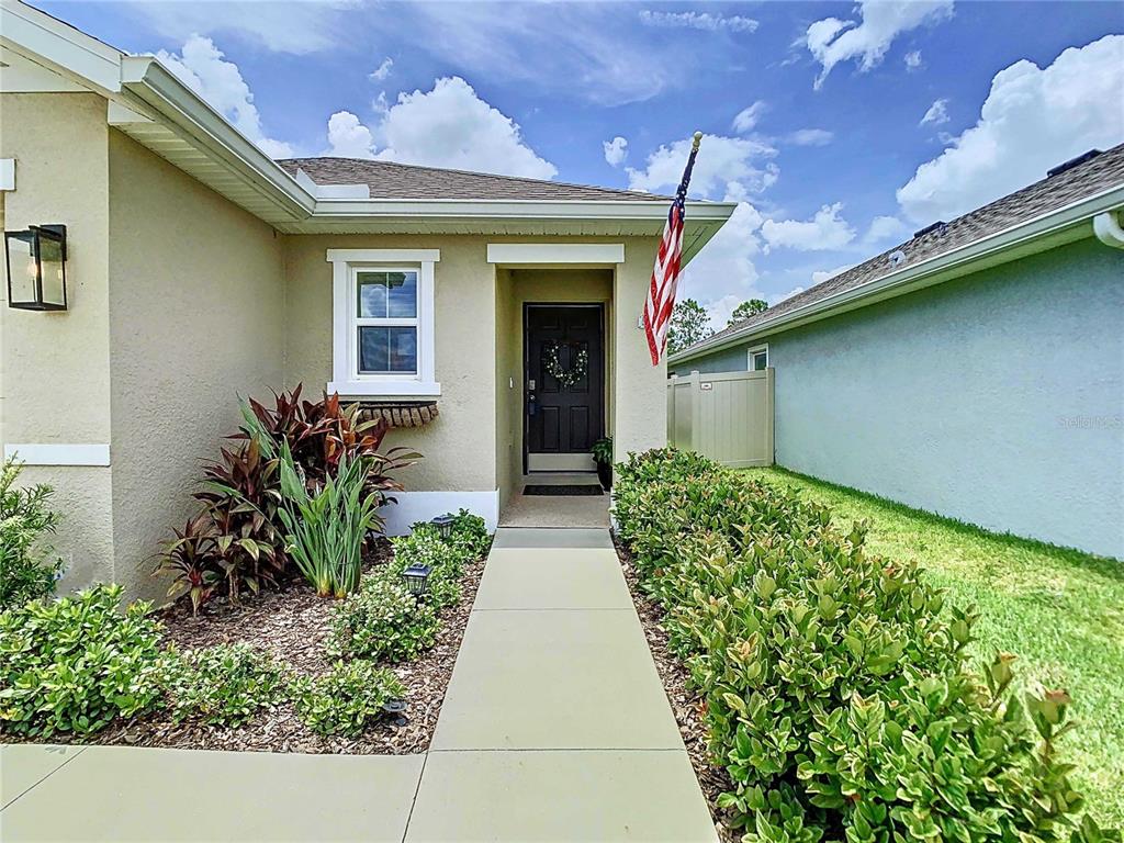 A concrete walkway leads to the front entrance of a house with tan siding — 7737 SAIL CLOVER LANE, ZEPHYRHILLS, FL