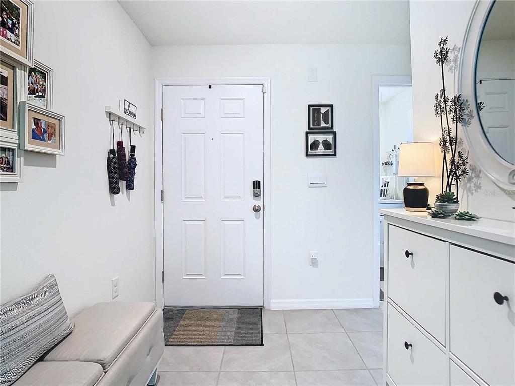 A bright entryway features a white door, tiled floors, a storage bench, and wall decor — 7737 SAIL CLOVER LANE, ZEPHYRHILLS, FL