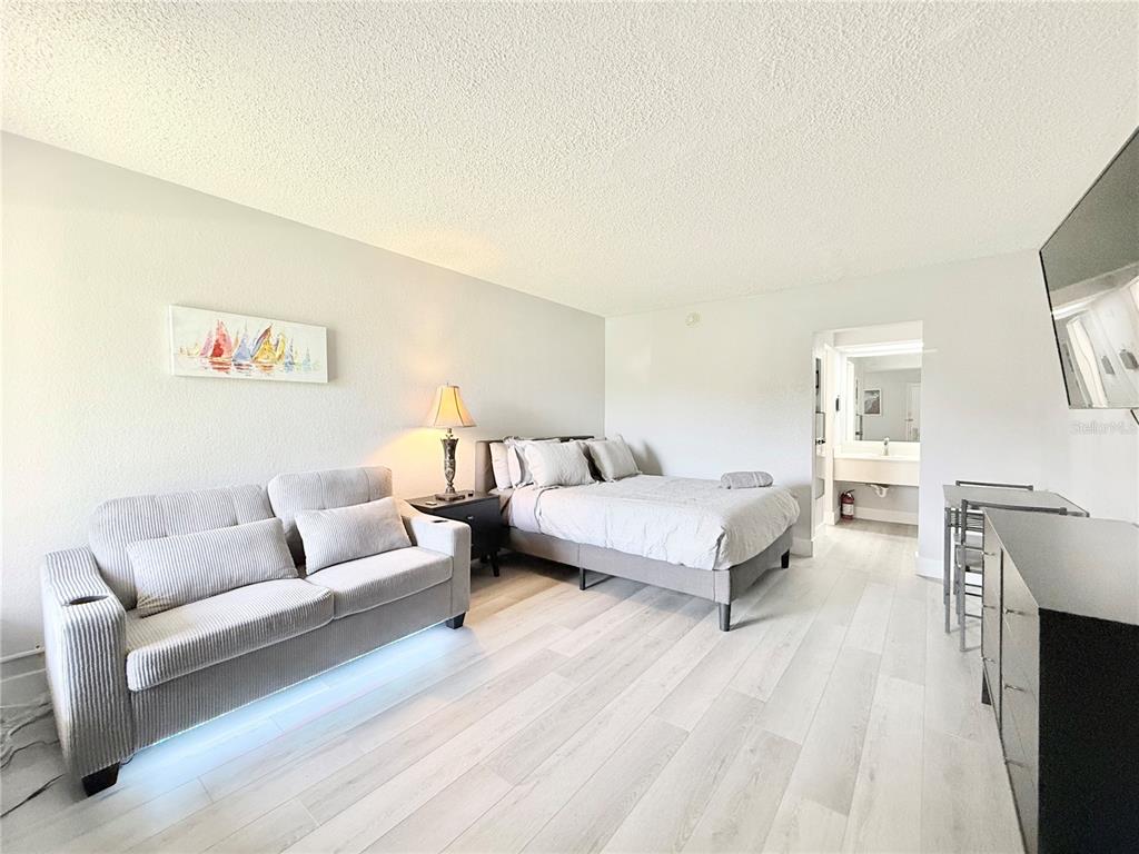 A studio apartment features a gray sofa, a bed, light wood flooring, and a bathroom entrance — 7150 N TAMIAMI TRAIL UNIT C126, SARASOTA, FL