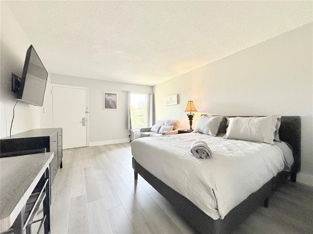 A bedroom features a bed, a television, a small sofa, and light-colored wood-look flooring — 7150 N TAMIAMI TRAIL UNIT C126, SARASOTA, FL