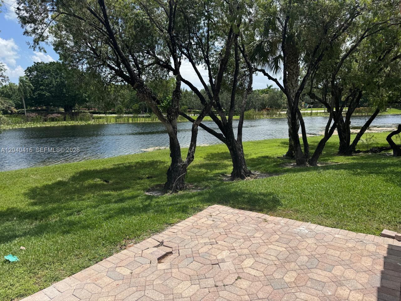 A paved patio overlooks a grassy lawn and a lake surrounded by mature trees — 2360 Dover, Weston FL 33326, WESTON, FL
