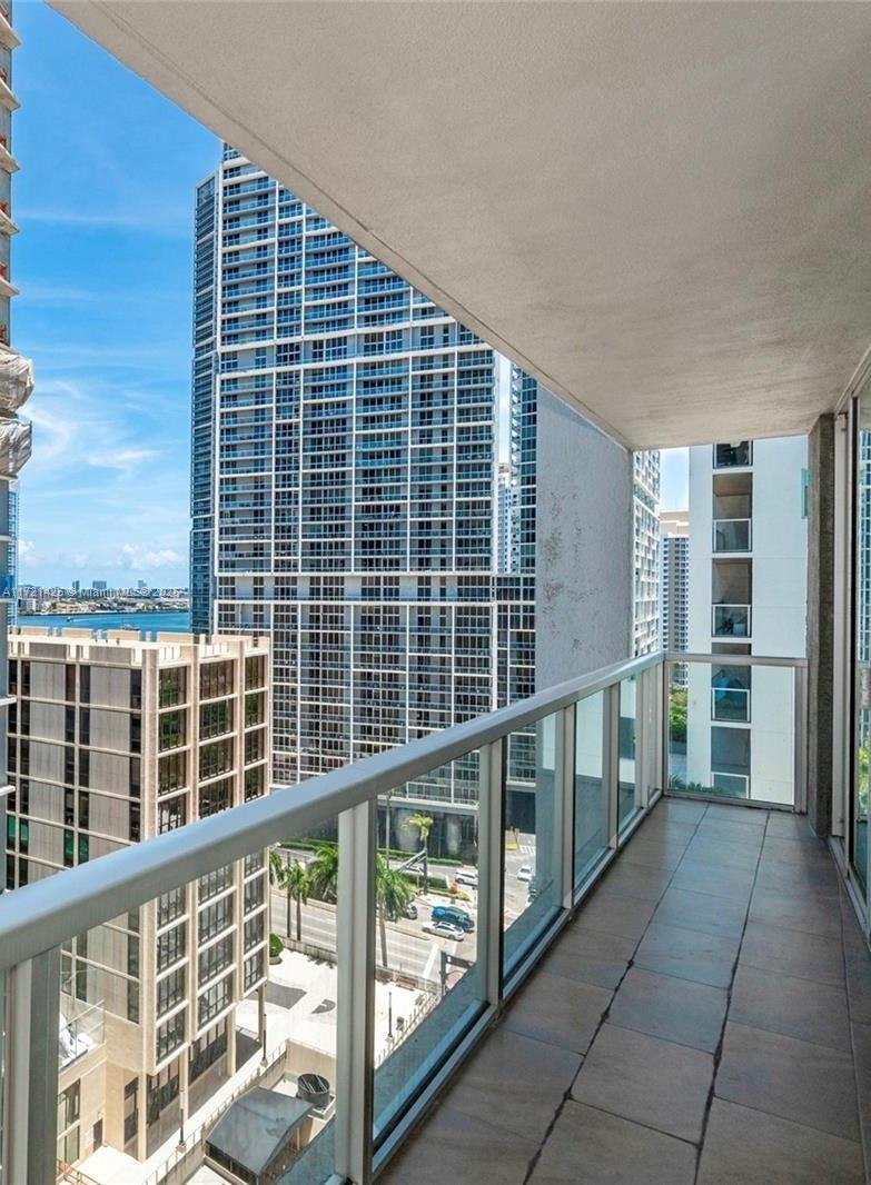 A high-rise balcony with glass railings overlooks city buildings and a distant body of water — 55 SE 6th St # 1507, Miami FL 33131, MIAMI, FL