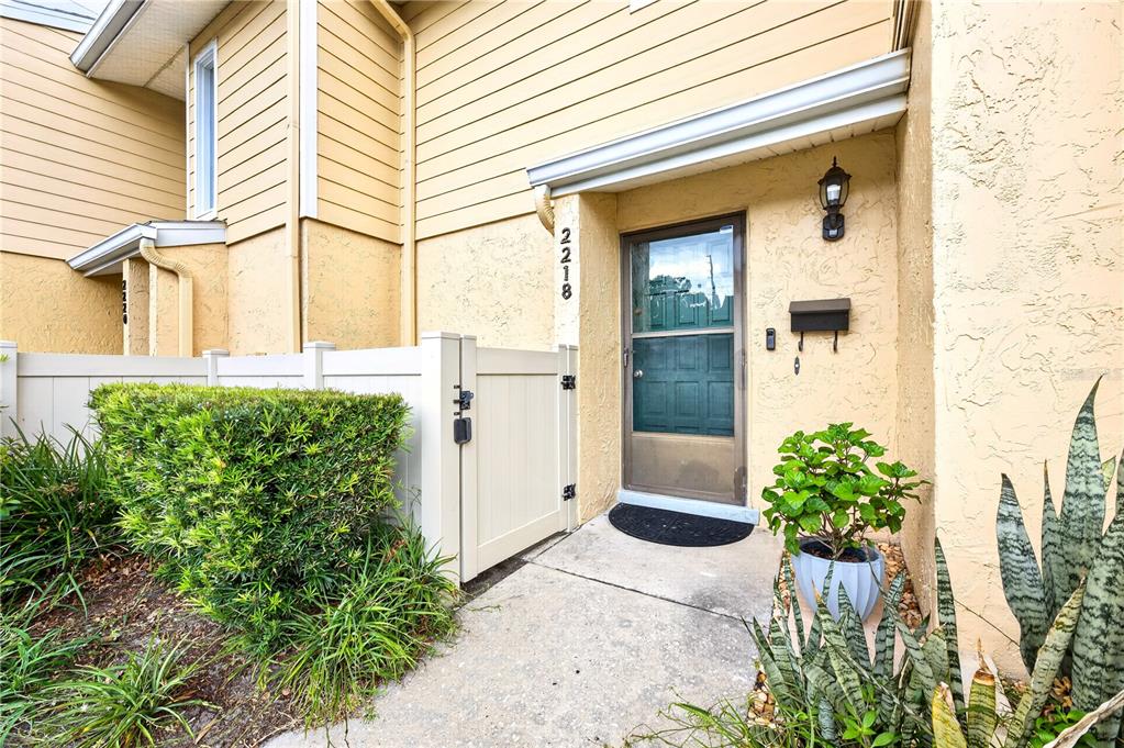 A tan townhouse entrance features a white vinyl fence, a green door, and potted plants — 2218 MCMAHON COURT UNIT 1038, ORLANDO, FL
