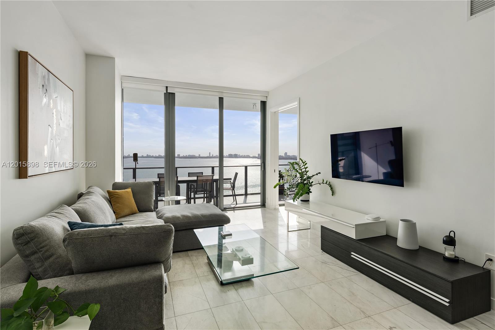 A modern living room features a grey sectional, glass coffee table, and balcony with water views — 480 NE 31st St # 1802, Miami FL 33137, MIAMI, FL