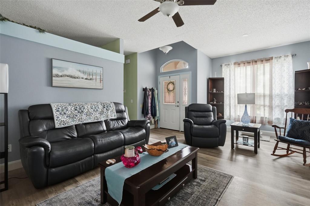 A living room features a black leather sofa, a recliner, a coffee table, and wood flooring — 1087 TRINIDAD AVENUE, SPRING HILL, FL