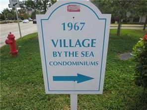 A white sign for Village by the Sea Condominiums stands on a grassy lawn — 1967 S Ocean Boulevard # D, Pompano Beach FL 33062, POMPANO BEACH, FL