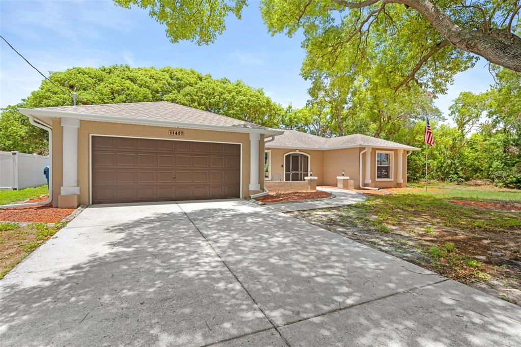 A tan single-story house with a two-car garage and a concrete driveway under large trees — 11487 KERRIDALE AVENUE, SPRING HILL, FL