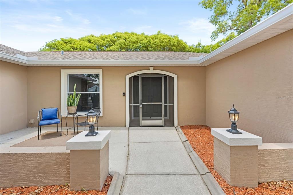 The exterior of a tan house features a concrete walkway leading to a screened front door — 11487 KERRIDALE AVENUE, SPRING HILL, FL