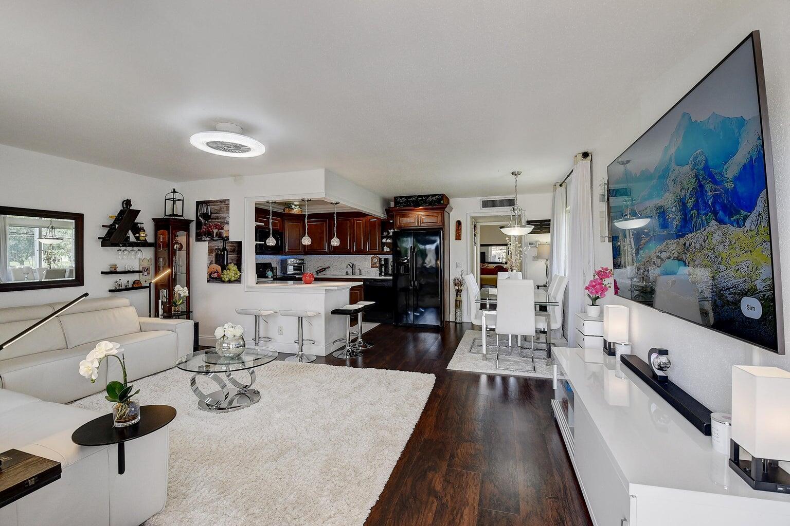 An open-concept living and dining area features dark wood floors, white furniture, and a kitchen — 123 Saxony C, Delray Beach FL 33446, DELRAY BEACH, FL