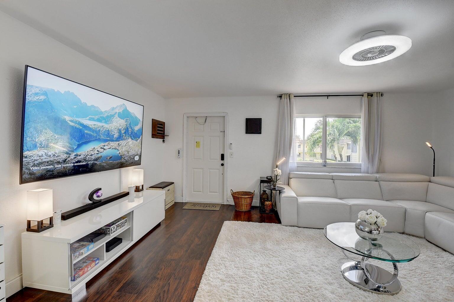 A living room with a large wall-mounted television, white sectional sofa, and dark wood flooring — 123 Saxony C, Delray Beach FL 33446, DELRAY BEACH, FL