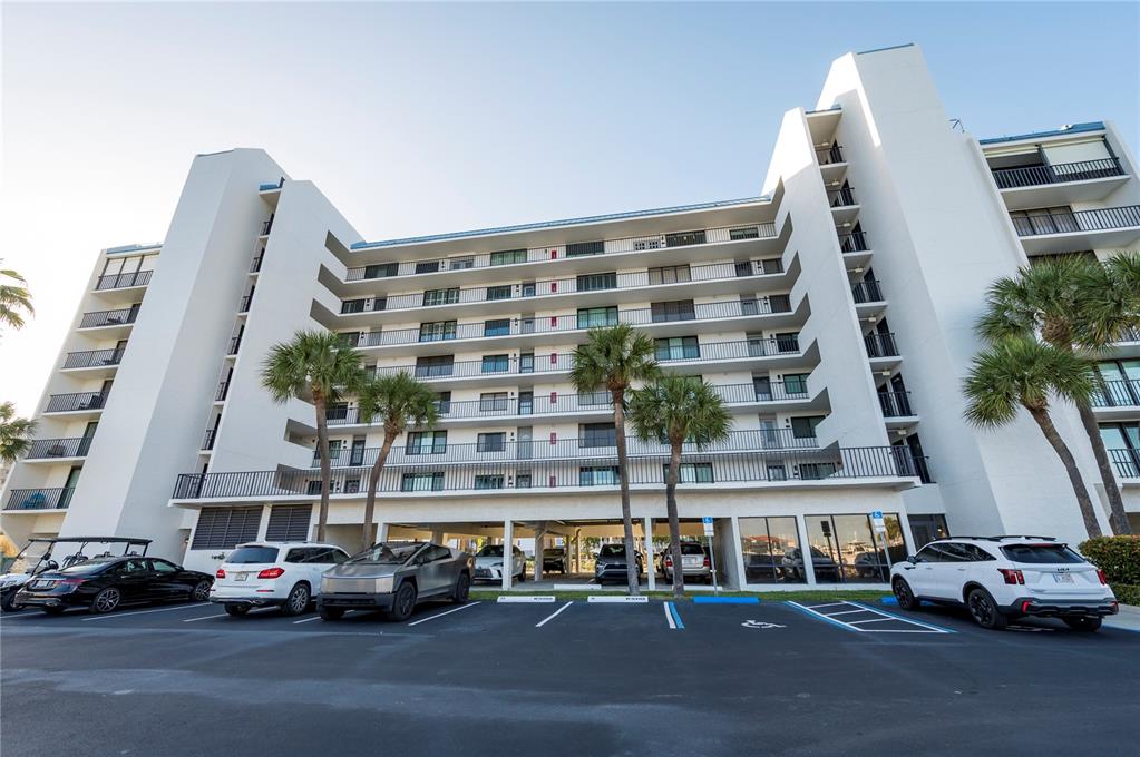 A low-angle exterior view of a multi-story white apartment building with a parking lot — 1591 GULF BOULEVARD UNIT 203S, CLEARWATER BEACH, FL