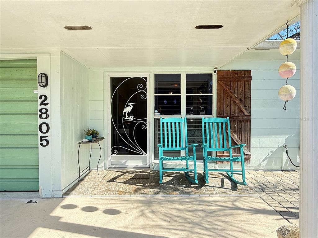 A covered porch features two turquoise rocking chairs, a decorative screen door, and house numbers — 2805 SAXON DRIVE, NEW SMYRNA BEACH, FL