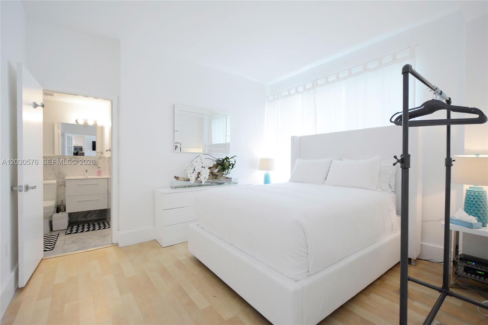 A bright bedroom features a white bed, light wood floors, and an attached bathroom — 1342 Drexel Ave # 105, Miami Beach FL 33139, MIAMI BEACH, FL