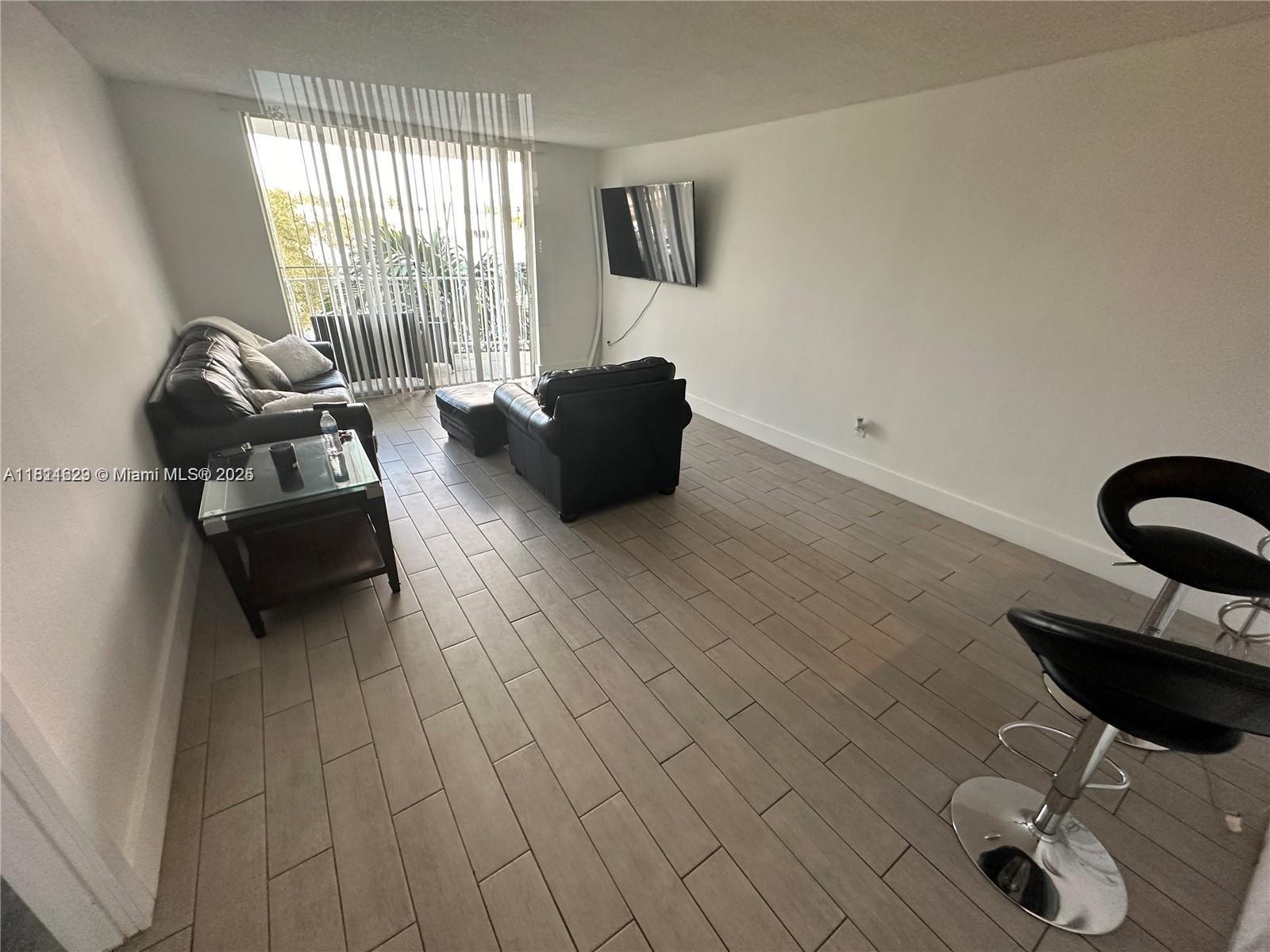 A living room features light wood-look tile flooring, black leather furniture, and a wall-mounted television — 486 NW 165th St Rd # 303, Miami FL 33169, MIAMI, FL