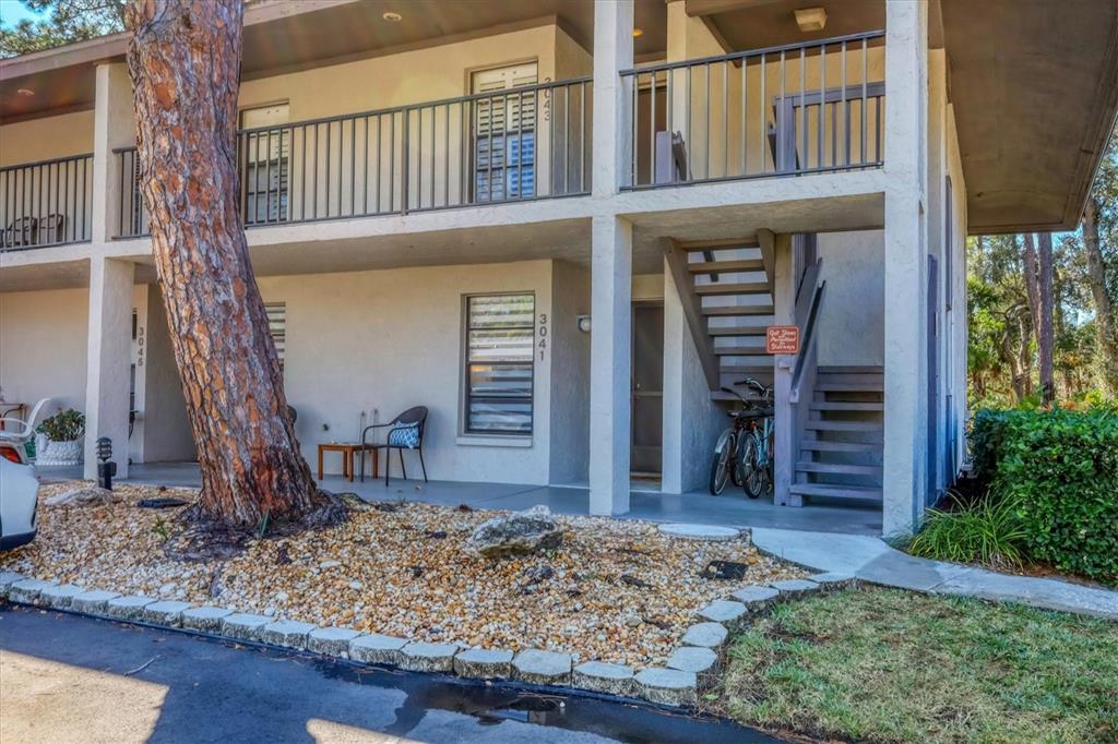 A two-story apartment building exterior features a staircase, a large tree, and gravel landscaping — 3041 QUAIL HOLLOW UNIT 27, SARASOTA, FL