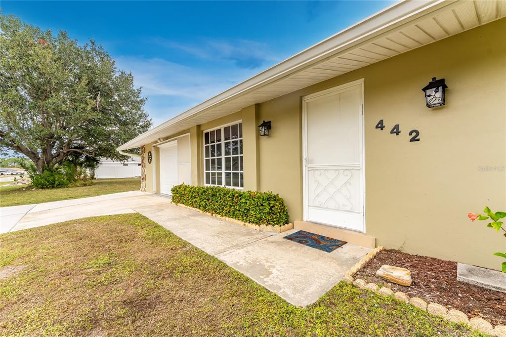 The exterior of a single-story house features a tan facade, a white door, and landscaping — 442 KOSTNER STREET, PORT CHARLOTTE, FL