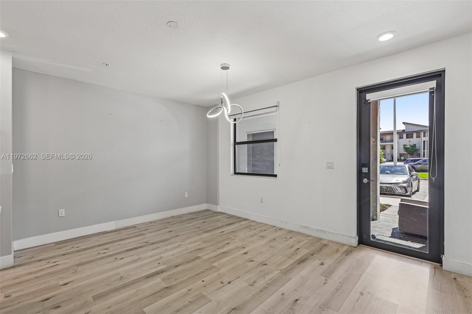 An empty room features light wood-look flooring, a modern light fixture, and a glass door — 10455 NW 82nd St # 4, Doral FL 33178, DORAL, FL