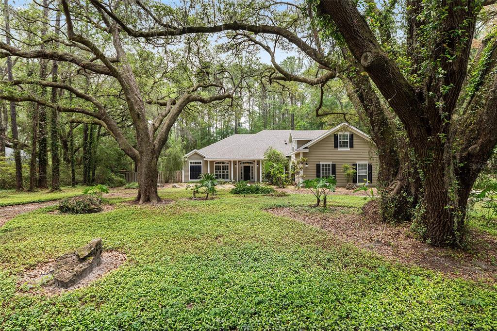 A tan house with black shutters sits behind large trees in a wooded yard — 10511 SW 17TH PLACE, GAINESVILLE, FL
