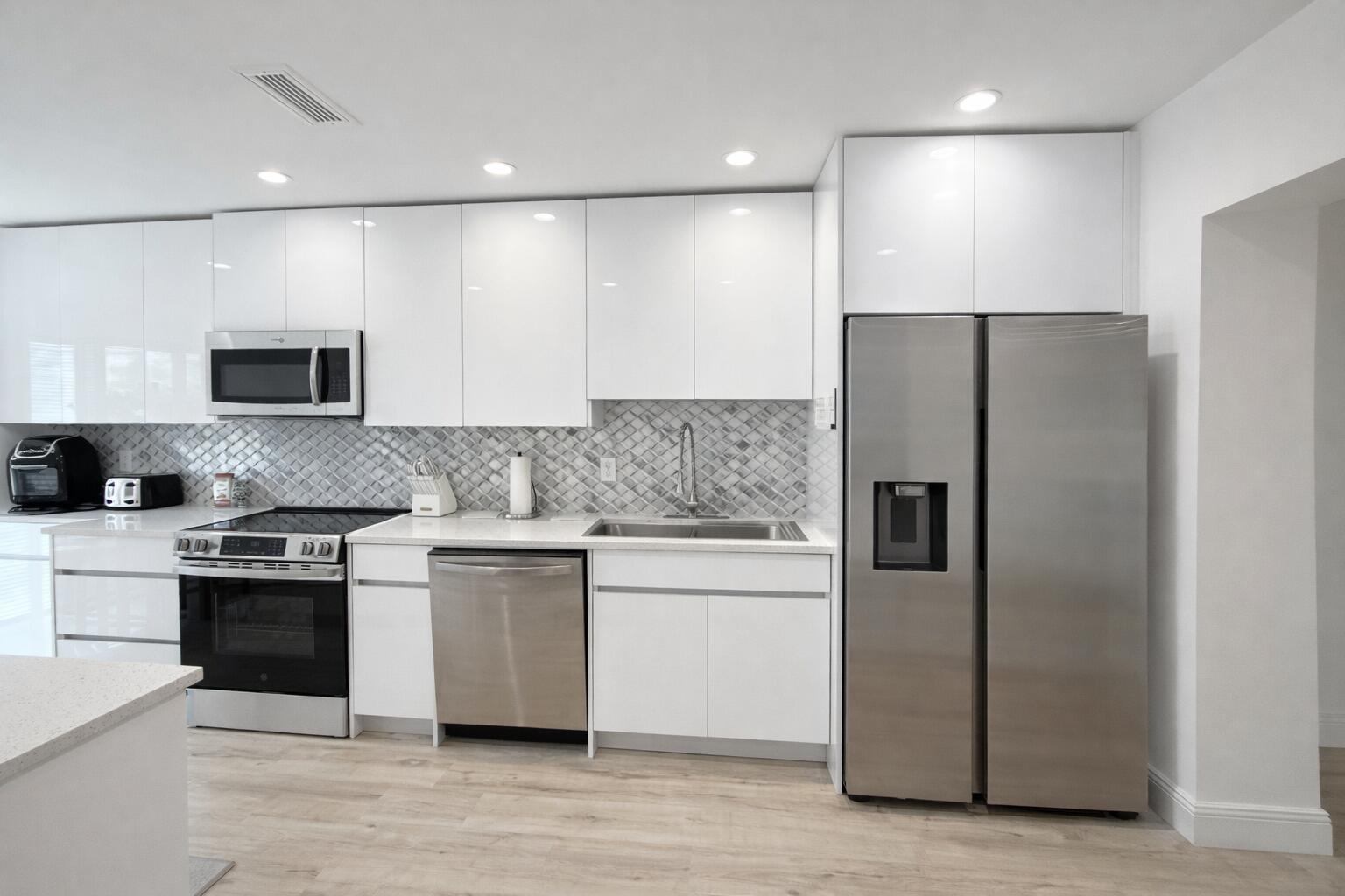 A modern kitchen features white high-gloss cabinets, stainless steel appliances, and a gray tiled backsplash — 4 Westgate Lane # 4c, Boynton Beach FL 33436, BOYNTON BEACH, FL