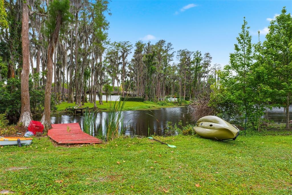 A grassy backyard features a small pond, a red wooden dock, and two kayaks — 19003 CEDAR LANE, LUTZ, FL