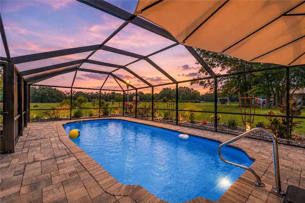 A screened-in swimming pool with a paver deck overlooks a grassy backyard at sunset — 11990 156TH STREET, MC ALPIN, FL