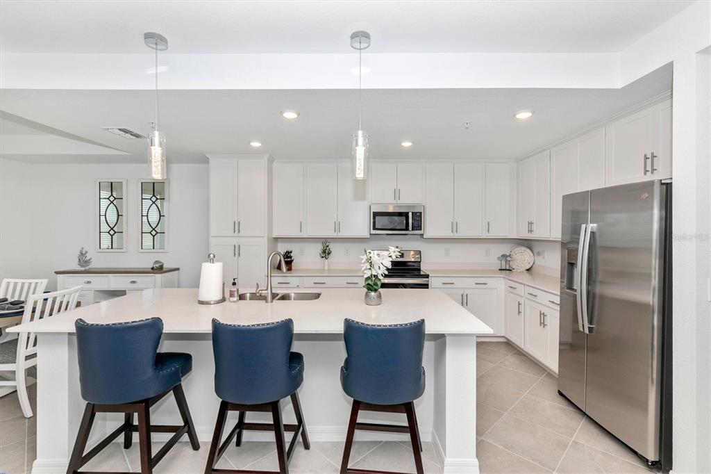 A modern kitchen features white cabinetry, a large island with blue stools, and stainless appliances — 12280 WELLEN GOLF STREET UNIT 203, VENICE, FL