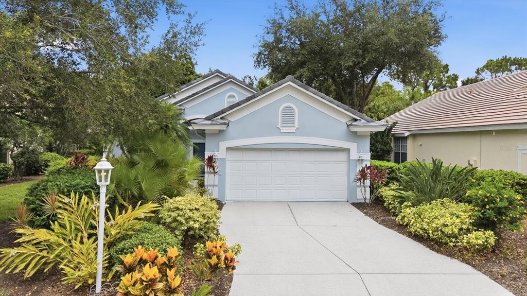 A light blue single-story house features a white garage door and lush tropical landscaping — 7147 PRESTWICK COURT, UNIVERSITY PARK, FL
