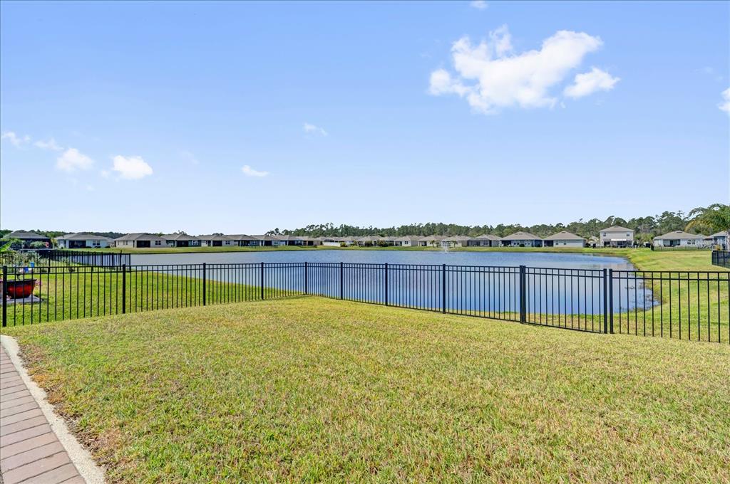 A backyard lawn overlooks a lake with a black metal fence and distant houses — 224 VENETIAN PALMS BOULEVARD, NEW SMYRNA BEACH, FL