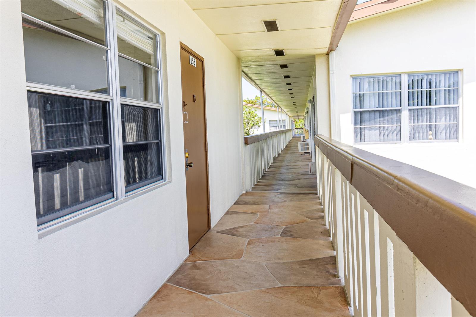 An outdoor apartment walkway features stone-patterned flooring, a brown door, and a white railing — 158 Mansfield D Drive, Boca Raton FL 33434, BOCA RATON, FL