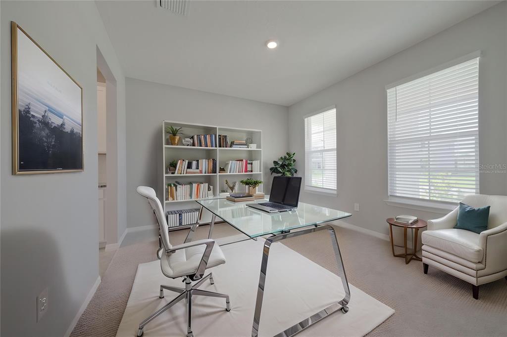A home office features a glass desk, white chair, bookshelf, and a window with blinds — 5748 WOODLAND SAGE DRIVE, SARASOTA, FL