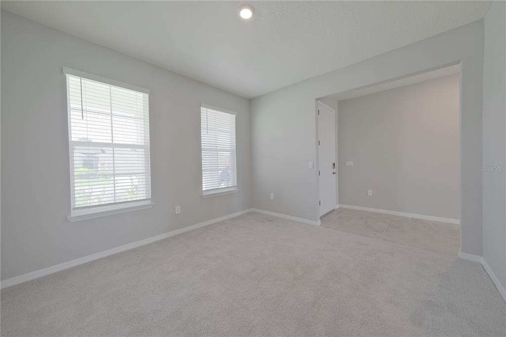 An empty room features gray walls, carpeted flooring, two windows with blinds, and a tiled entryway — 5748 WOODLAND SAGE DRIVE, SARASOTA, FL