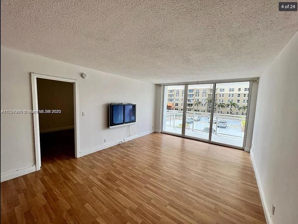 An empty living room features wood-look flooring, a wall-mounted television, and a sliding glass balcony door — 2030 S Ocean Dr # 211, Hallandale Beach FL 33009, HALLANDALE BEACH, FL