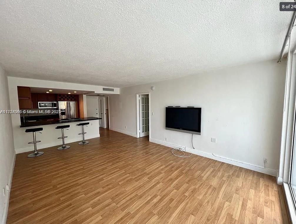 A living room with wood-look flooring features a wall-mounted television and an open kitchen — 2030 S Ocean Dr # 211, Hallandale Beach FL 33009, HALLANDALE BEACH, FL