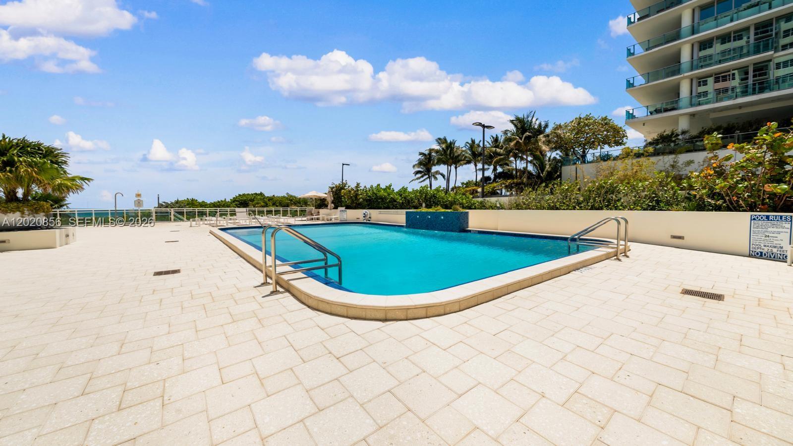 A rectangular swimming pool with a tiled deck is surrounded by landscaping and a building — 6917 Collins Ave # 107, Miami Beach FL 33141, MIAMI BEACH, FL