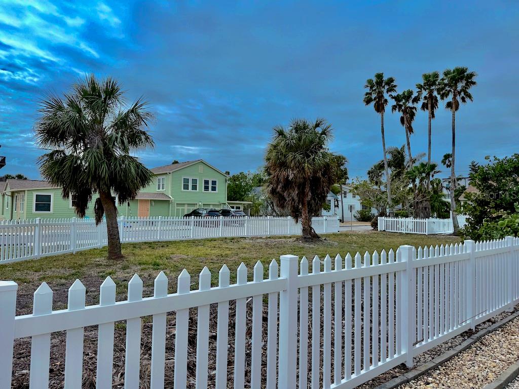 A fenced-in vacant lot features several palm trees with a light green house in the background — 1300 PASS A GRILLE WAY, ST PETE BEACH, FL