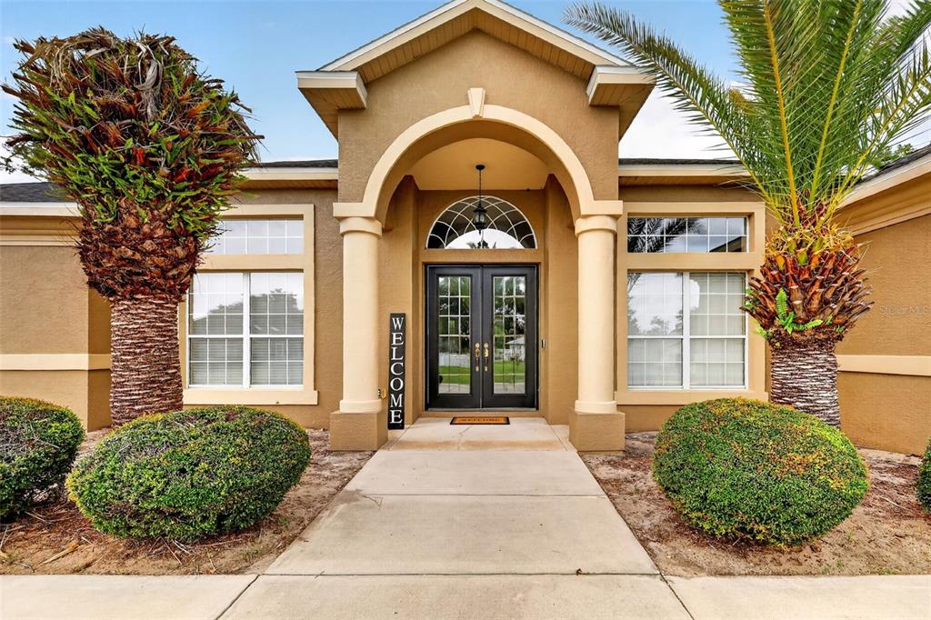 The front exterior of a house features a tan stucco facade, double glass doors, and two palm trees — 485 NW 45TH LANE, OCALA, FL