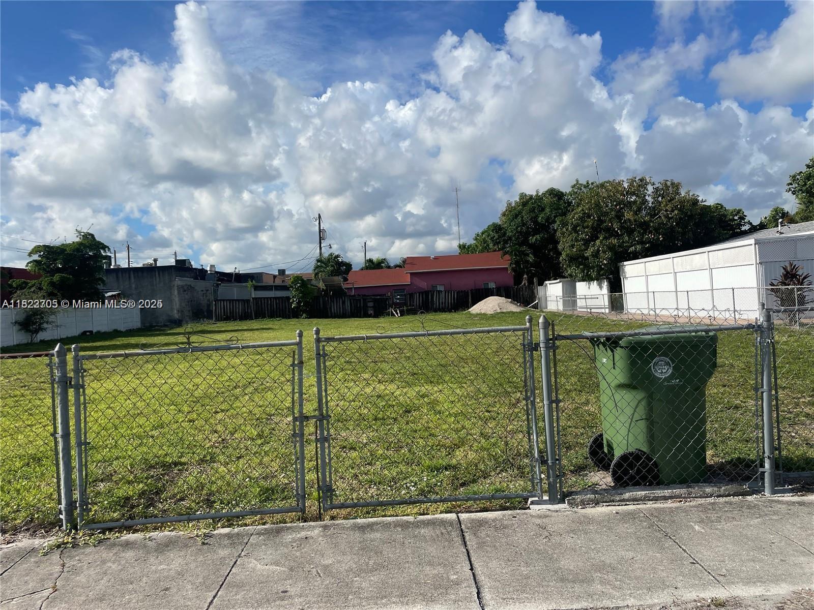 A fenced, vacant grassy lot sits under a cloudy blue sky — 20 W 40th Pl, Hialeah FL 33012, HIALEAH, FL
