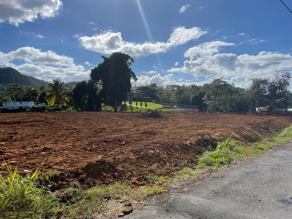 A large, cleared dirt lot sits beside a paved road under a blue, cloudy sky — BO. QUEBRADA NEGRITO, TRUJILLO ALTO, PR