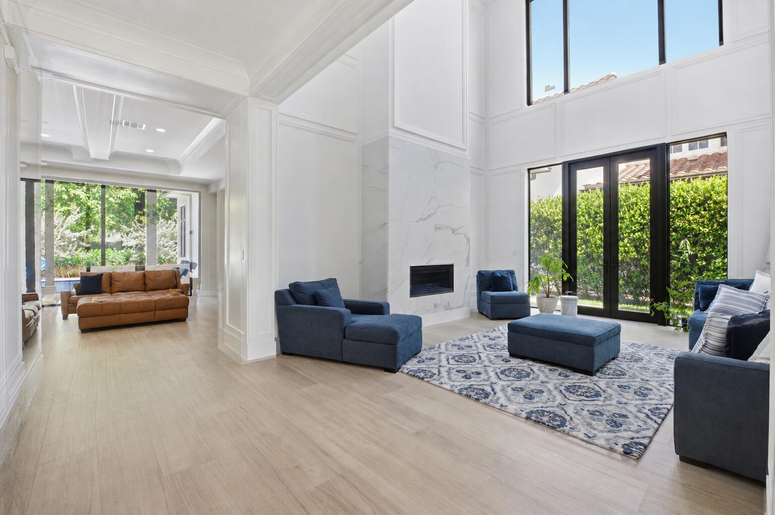 A spacious living room features a marble fireplace, blue furniture, and large windows overlooking greenery — 16075 Tuscany Estates Drive, Delray Beach FL 33446, DELRAY BEACH, FL