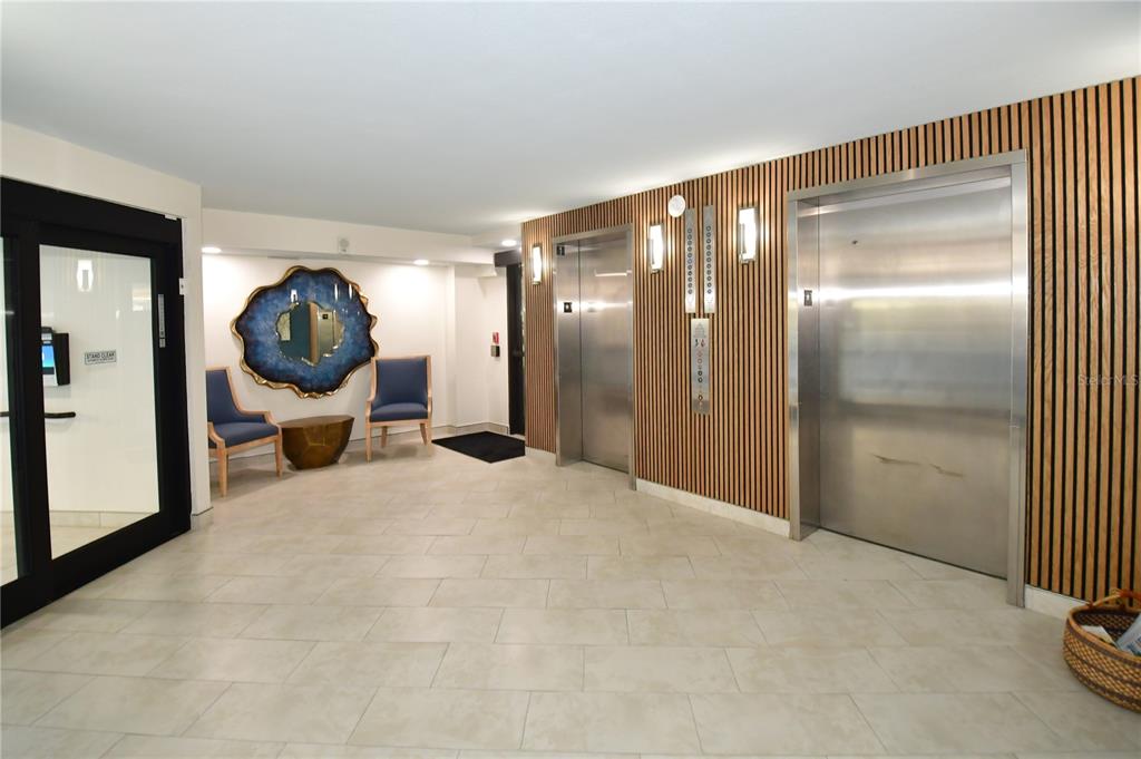 A lobby features two stainless steel elevators, wood slat wall paneling, and two blue chairs — 690 ISLAND WAY UNIT 208, CLEARWATER, FL