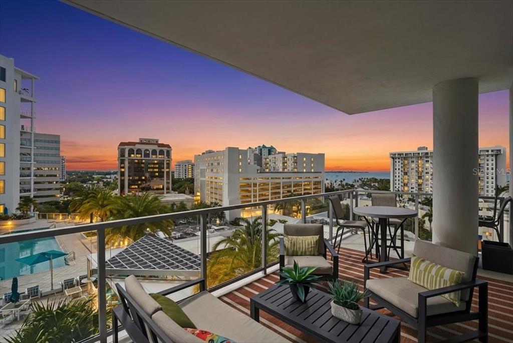 A balcony with outdoor furniture overlooks a swimming pool and a city skyline at sunset — 111 S PINEAPPLE AVENUE UNIT 714, SARASOTA, FL