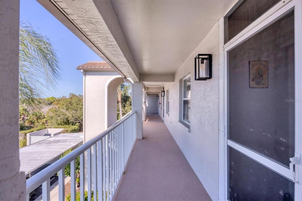 An outdoor covered walkway features a white railing, beige flooring, and a screen door — 8735 OLDE HICKORY AVENUE UNIT 8303, SARASOTA, FL