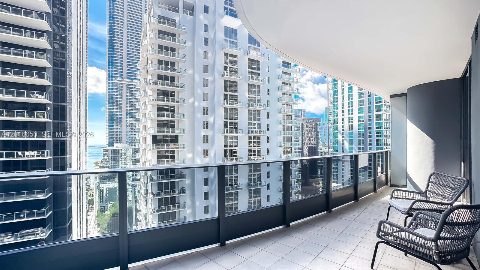 A high-rise balcony features a tiled floor, glass railings, and two dark wicker chairs — 1000 Brickell Plz # 2603, Miami FL 33131, MIAMI, FL